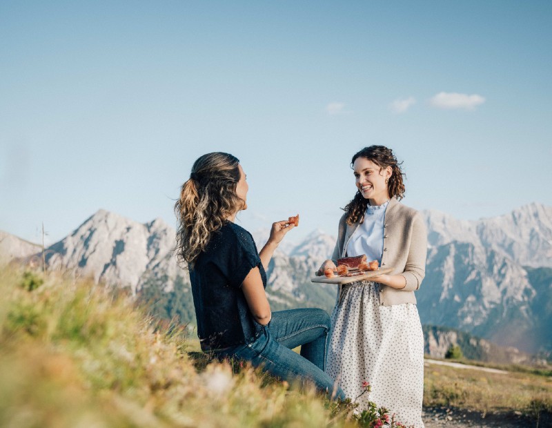 Zwei Frauen unterhalten sich lachend auf einer Wiese mit Bergblick, eine hält einen Teller mit aufgeschnittenem Speck.