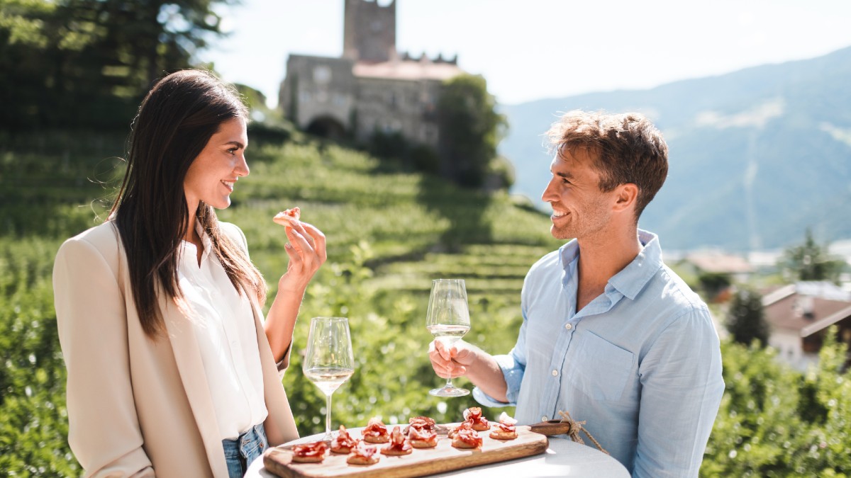 Una coppia è seduta fuori a un tavolino con un piatto di pancetta e due bicchieri di vino.