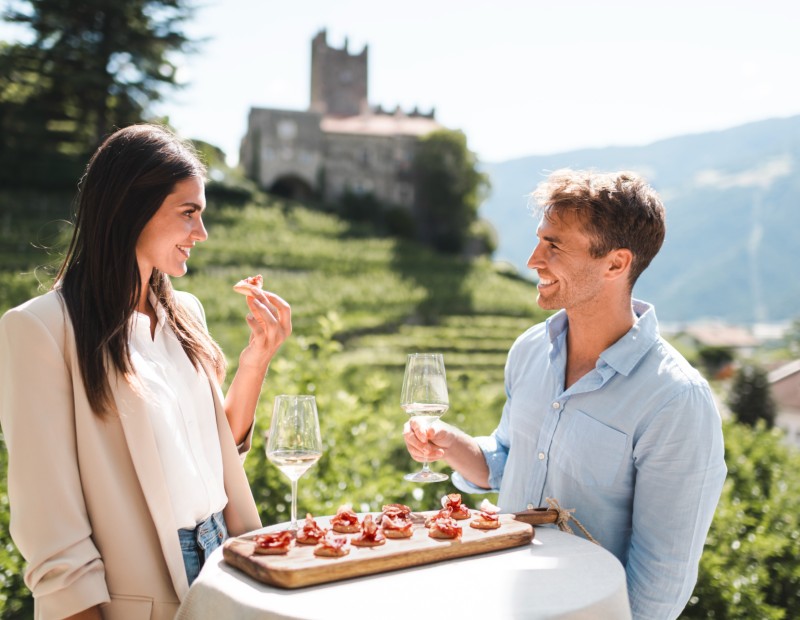Ein Paar sitz im Freien an einem kleinen Tisch mit einer Platte Speck und 2 Gläsern Wein.