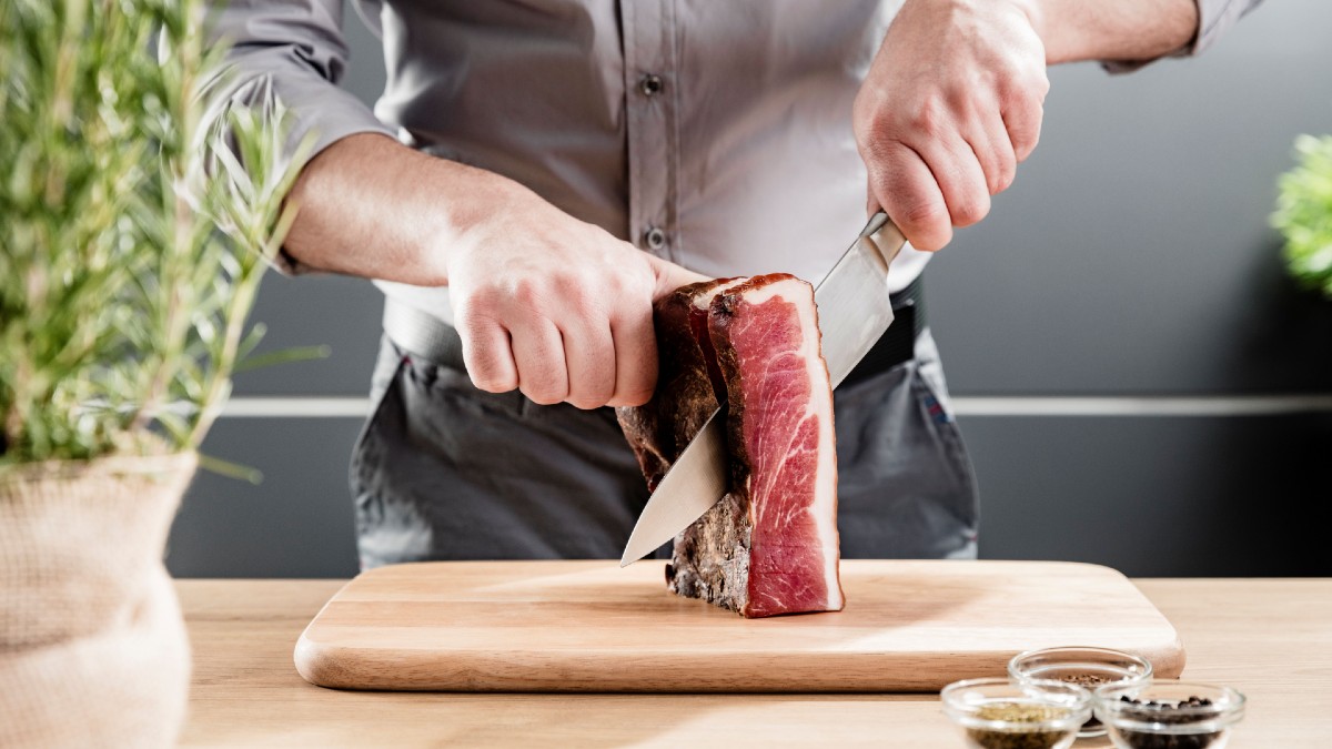 Ein Mann zeigt wie man den Südtioler Speck g.g.A. richtig mit der Hand schneidet