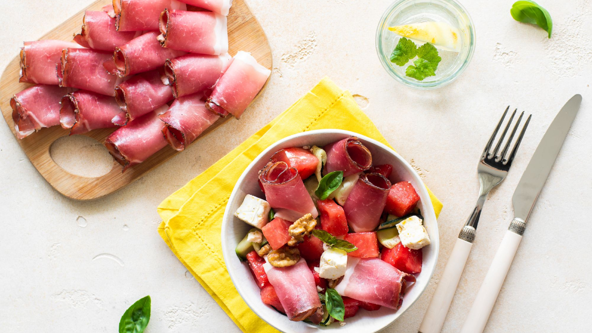 Insalata di anguria, feta e Speck Alto Adige IGP in una ciotola con tovagliolo giallo e bicchiere accanto.
