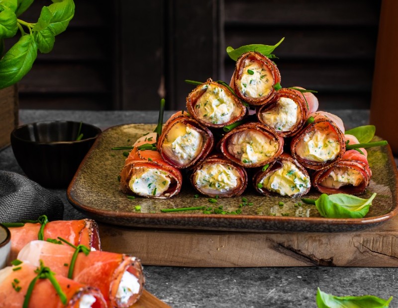 Röllchen aus Südtiroler Speck g.g.A. mit Kräuter-Frischkäse-Füllung, angerichtet auf einem Teller mit frischen Kräutern.
