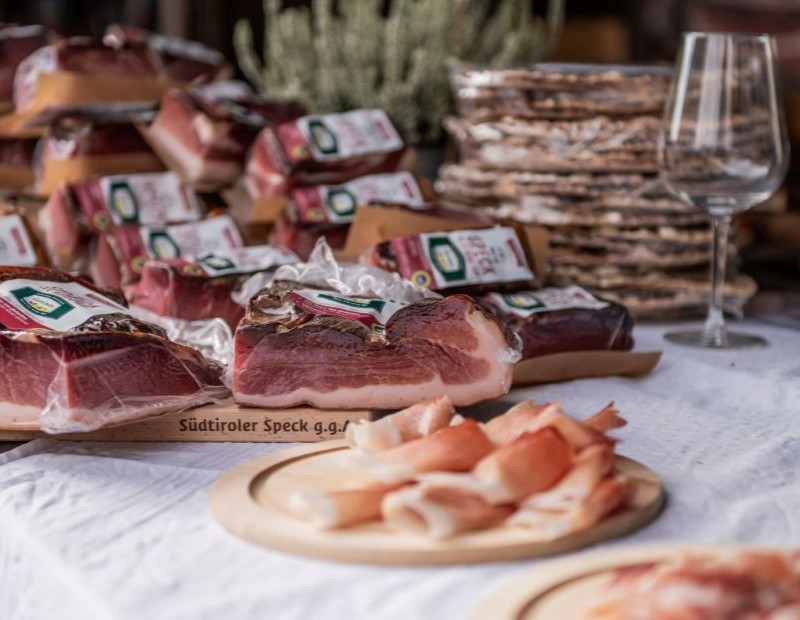 Aufgeschnittener Südtiroler Speck g.g.A. auf Holztellern, daneben verpackte Speckstücke und ein Weinglas auf einem gedeckten Tisch.