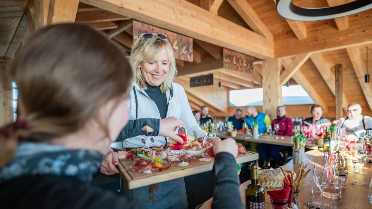 Eine Frau serviert eine Speckplatte mit Südtiroler Speck g.g.A. und Beilagen in einer Berghütte, im Hintergrund Gäste an langen Tischen.