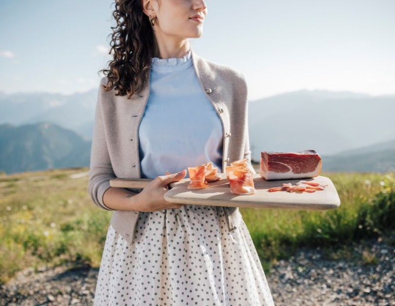 Donna in abito tradizionale tiene in mano un tagliere di legno con fettine sottili di Speck Alto Adige su un prato alpino. Sullo sfondo, le Dolomiti risplendono sotto un cielo terso.