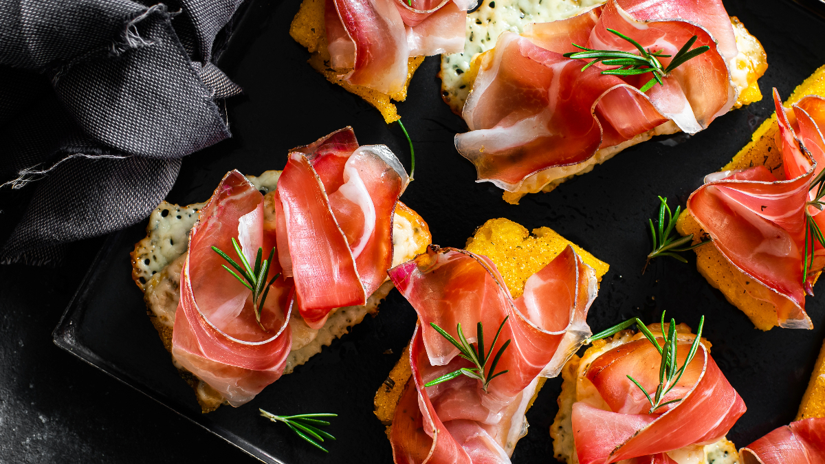 Gegrillte Polentascheiben mit geschmolzenem Käse, belegt mit Speck und Rosmarinzweigen, serviert auf einem schwarzen Teller.