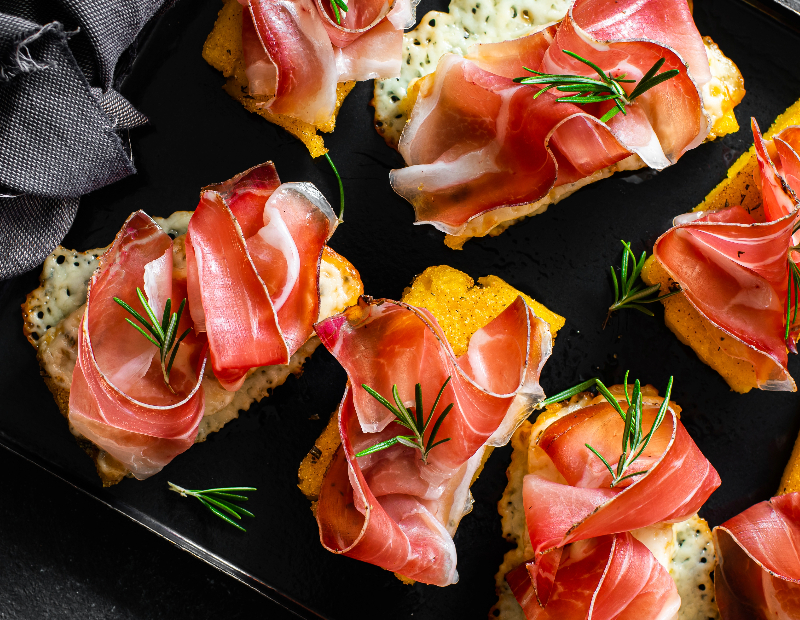 Gegrillte Polentascheiben mit geschmolzenem Käse, belegt mit Speck und Rosmarinzweigen, serviert auf einem schwarzen Teller.