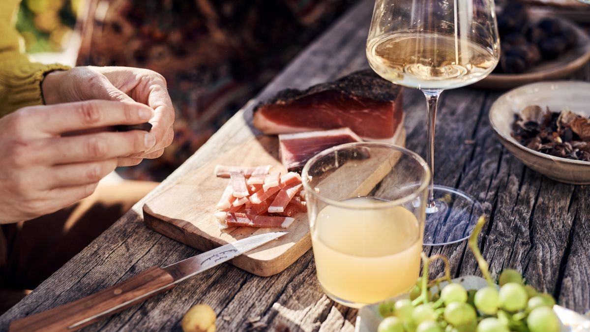 Una persona affetta lo speck su un tavolo rustico all’aperto. Accanto: fettine di Speck Alto Adige, uva, castagne e bicchieri di vino e succo di mela – una tipica scena autunnale di Törggelen.