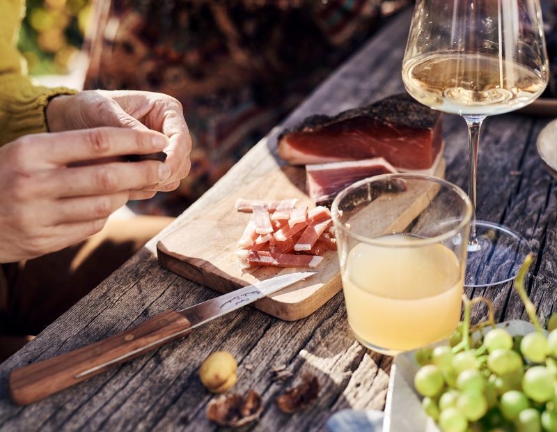 Person bereitet Speck auf einem Holztisch zu, daneben liegen geschnittener Südtiroler Speck, Trauben, Kastanien und Gläser mit Wein und Apfelsaft – typische Törggele-Szene im Freien.