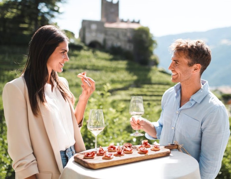 Ein Paar sitz im Freien an einem kleinen Tisch mit einer Platte Speck und 2 Gläsern Wein.
