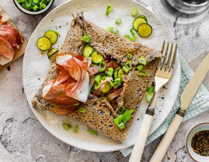 Galette bretonne servita su un piatto con zucchine, cipollotto e Speck Alto Adige IGP, decorata con fette fresche di speck e pepe nero.