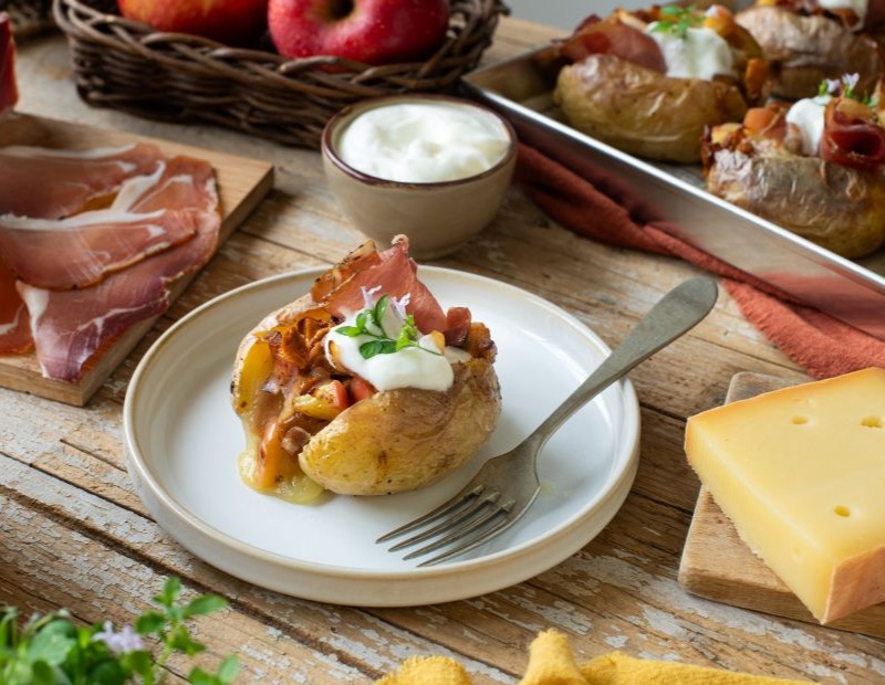 Una patata al forno con Speck Alto Adige IGP, finferli, mela, fontina e panna acida su un piatto. Intorno si vedono gli ingredienti e la teglia con altre patate.