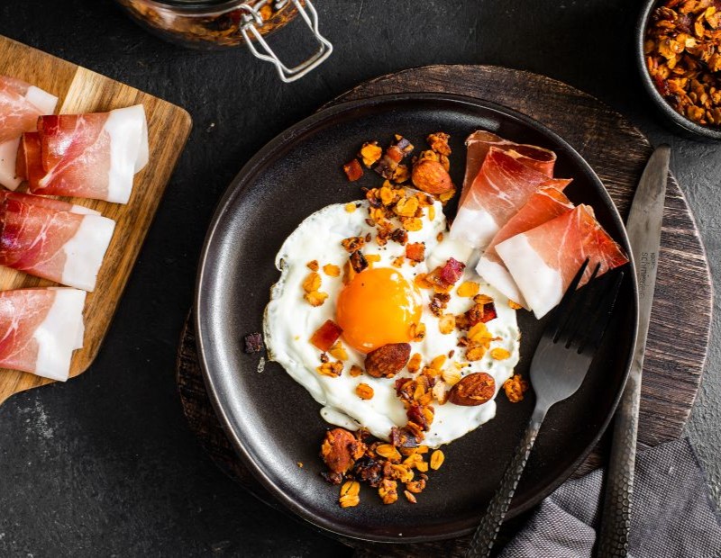 Spiegelei auf einem dunklen Teller mit knuspriger Granola und Scheiben von Südtiroler Speck g.g.A., daneben eingerollte Speckscheiben auf einem Holzbrett.