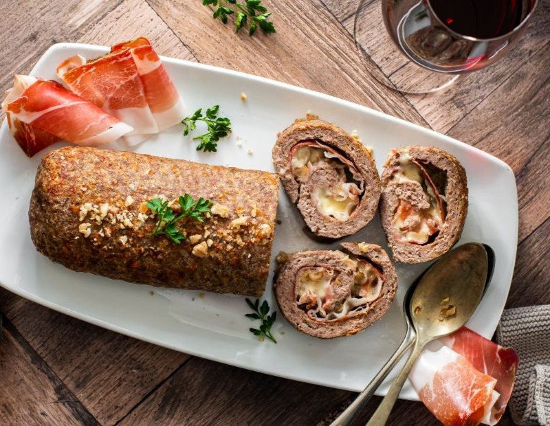 Sliced stuffed meatloaf on a white plate, with melted cheese and speck visible in each slice, garnished with fresh parsley and served with thin slices of speck and a glass of red wine.