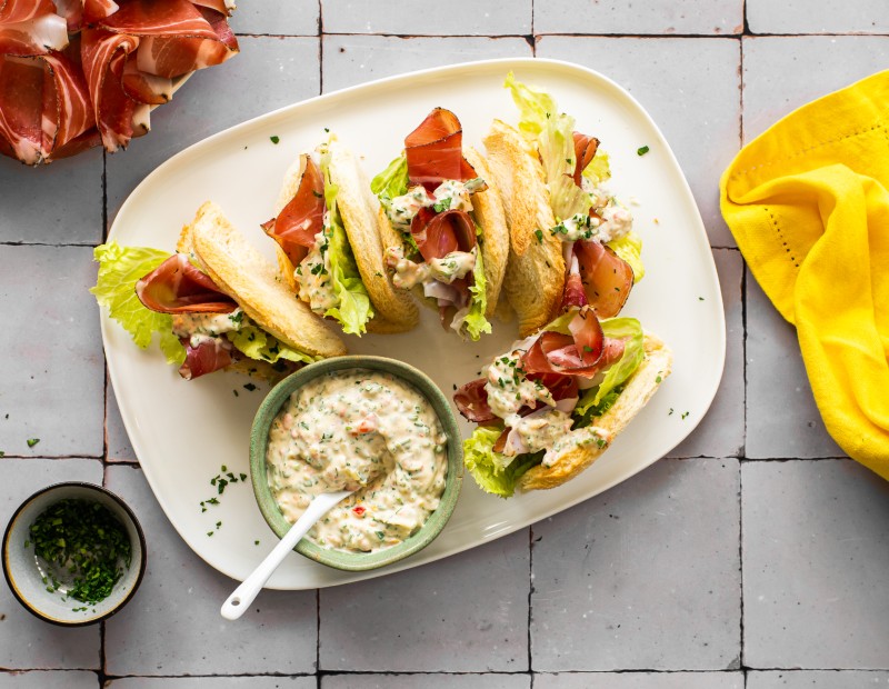 Geröstete Sandwichhälften gefüllt mit Salat, Speck und cremiger Kräutersauce, angerichtet auf einem weißen Teller mit einem Schälchen Sauce daneben.