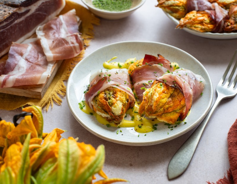 Tre fiori di zucca ripieni di ricotta e avvolti nello speck, serviti su un piatto con salsa cremosa e erbe fresche.
