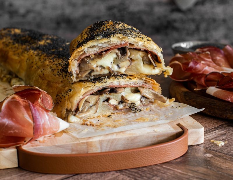 Sliced savoury strudel filled with mushrooms, Speck Alto Adige PGI and melted cheese, served on a wooden board with slices of speck on the side.