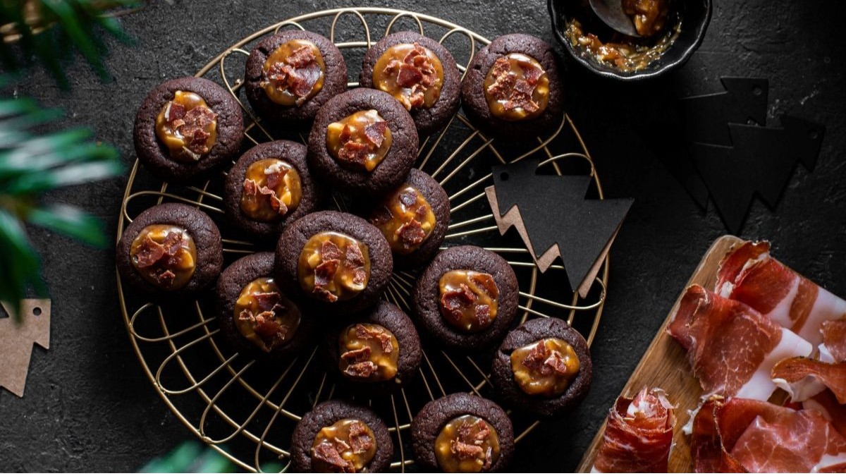 Mehrere Schokoladenkekse mit einer Füllung aus goldenem Erdnusskaramell und kleinen Speckstücken auf einem goldenen Kuchengitter angerichtet.
