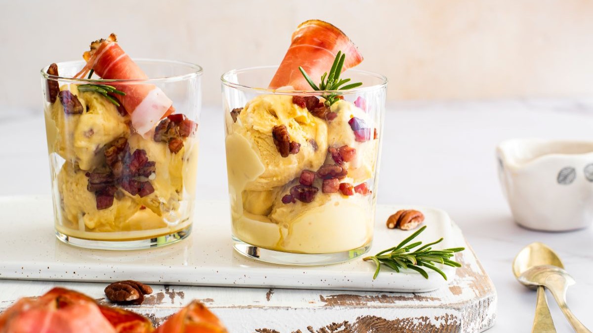 Zwei Dessertgläser gefüllt mit cremigem Speiseeis, garniert mit Speckwürfeln, Speckscheiben und einem Hauch Rosmarin.