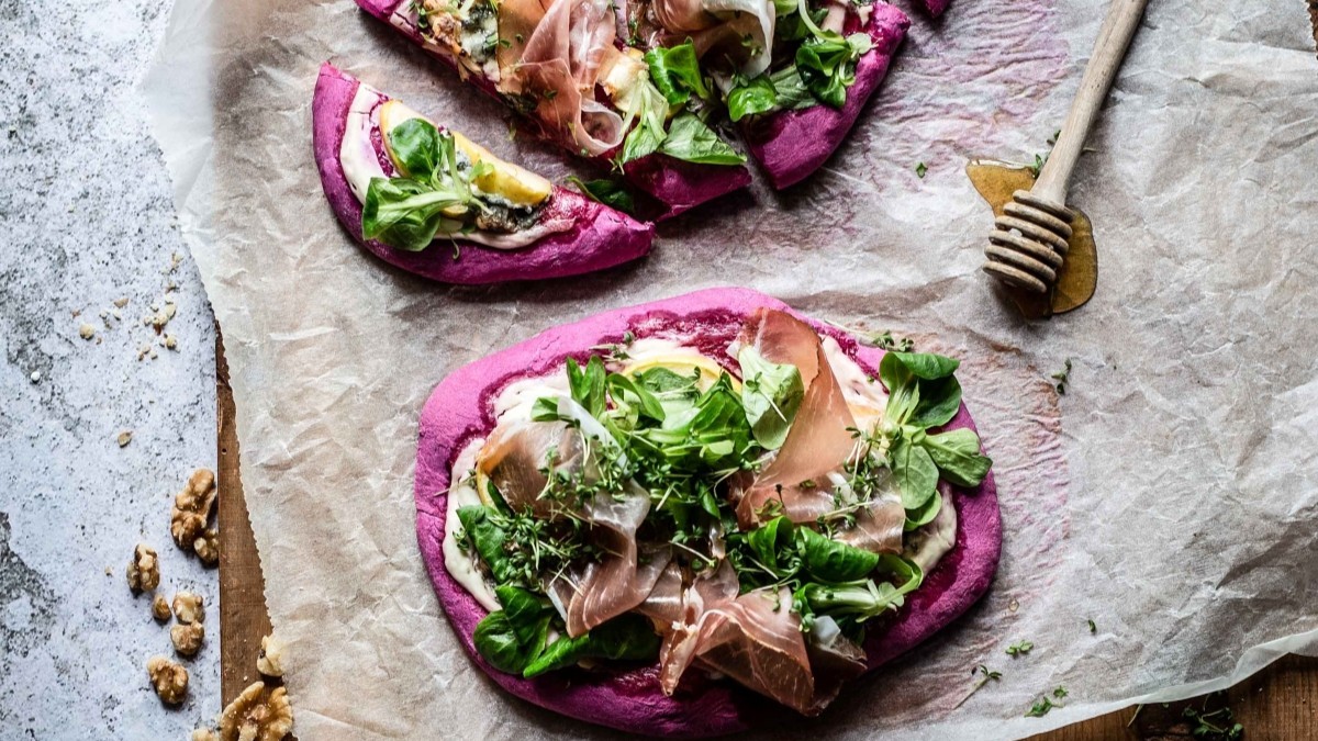 Knusprige, leuchtend rote Pizza aus Rote-Beete-Teig, belegt mit frischen Salatblättern, Südtiroler Speckscheiben, Apfelstücken und Gorgonzola, auf Backpapier angerichtet.