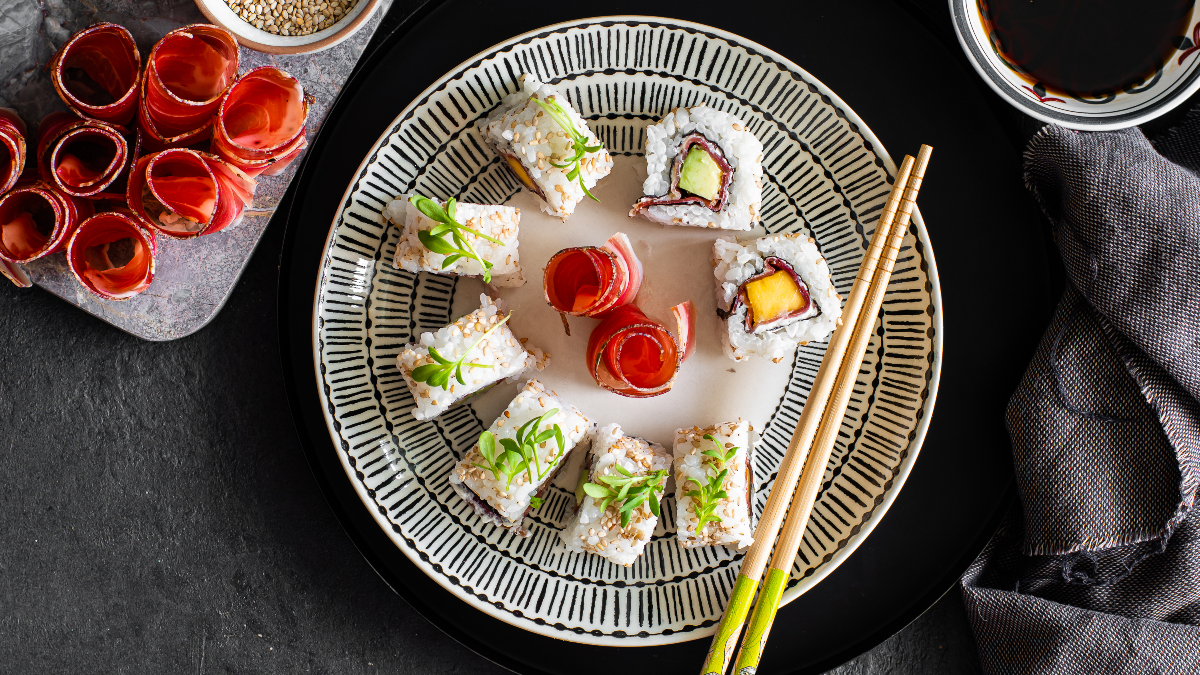 Uramaki-Sushi mit Speck, Mango und Avocado, angerichtet auf einem dekorativen Teller mit Stäbchen, serviert mit einem Schälchen Sojasauce.